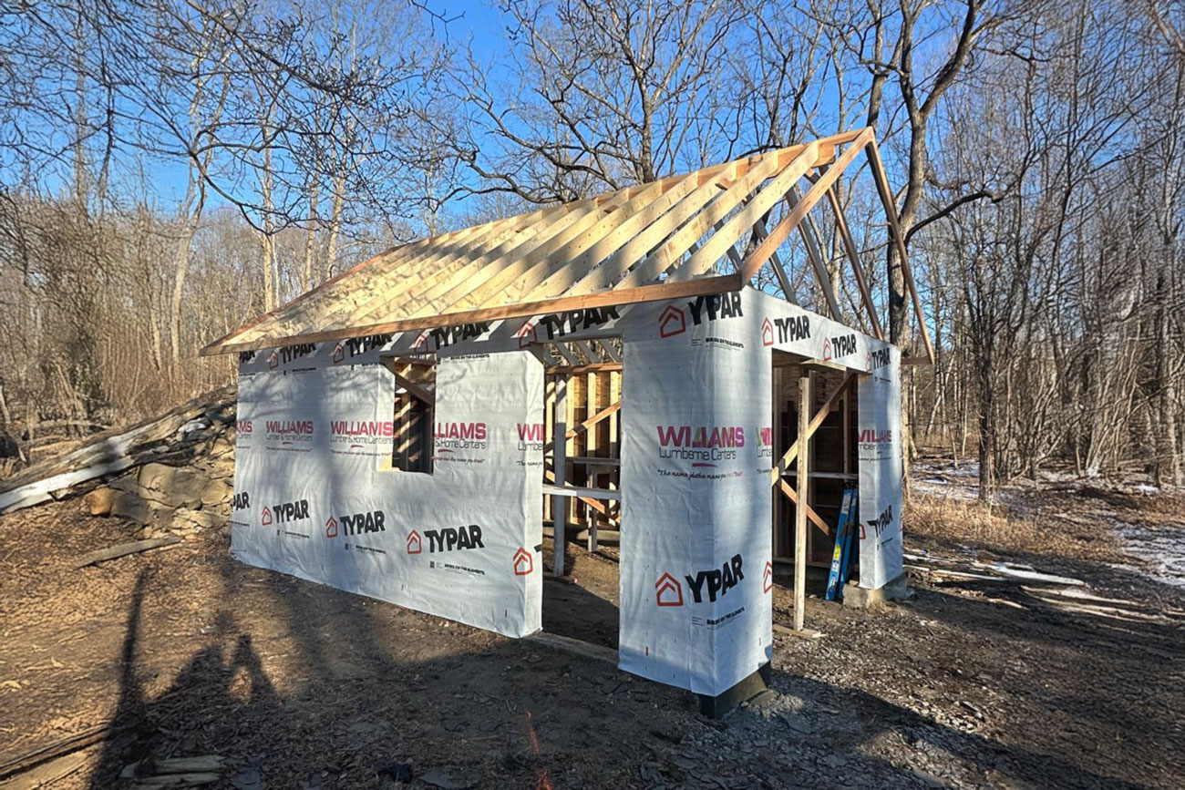 Construction site showing the framing of a separate garage with a roof under construction, surrounded by trees and a fire burning in a barrel.